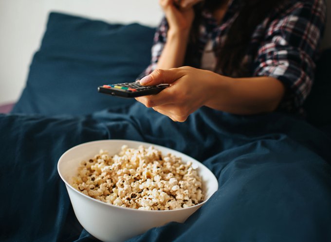 A person sitting on a couch holding a TV remote with a bowl of popcorn